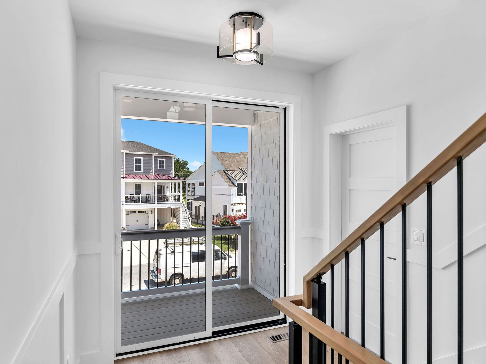 Stairwell with a balcony view of houses. White walls, black railing, and light wood steps. Bright blue sky.
