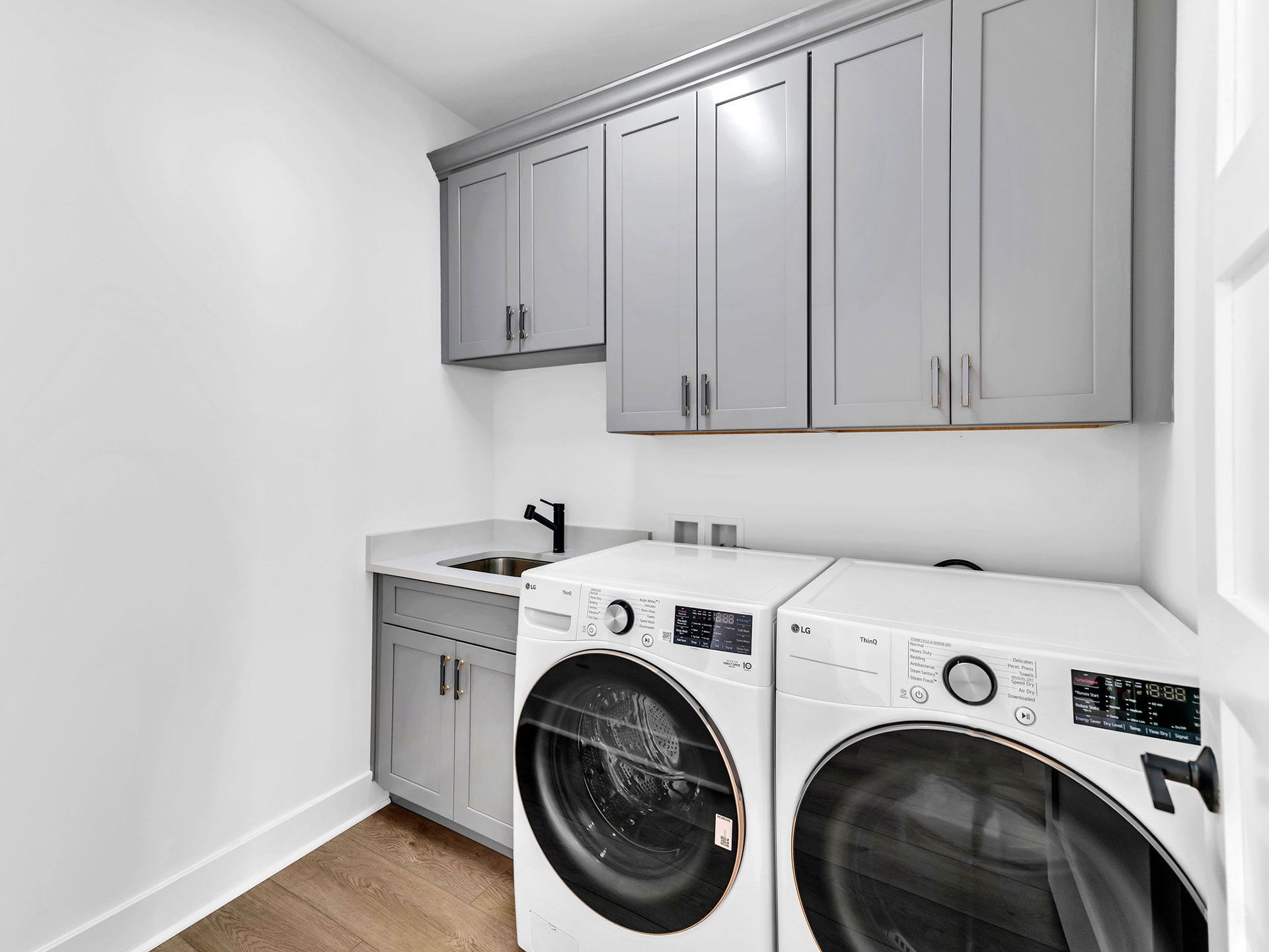 Laundry room with grey cabinets, white appliances, sink, and hardwood floor.