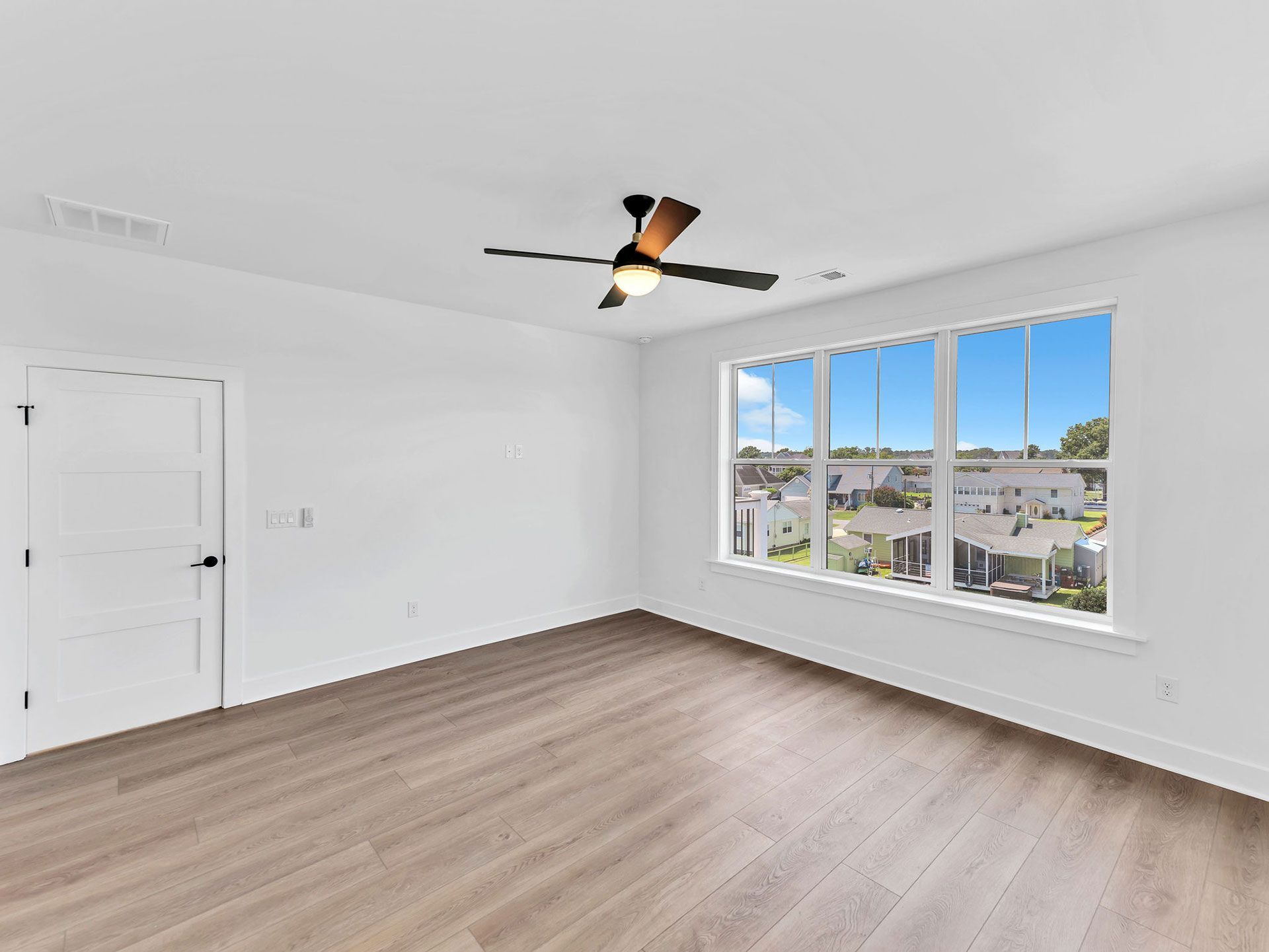 Empty, sunlit room with hardwood floors, white walls, and a ceiling fan near a large window overlooking a neighborhood.