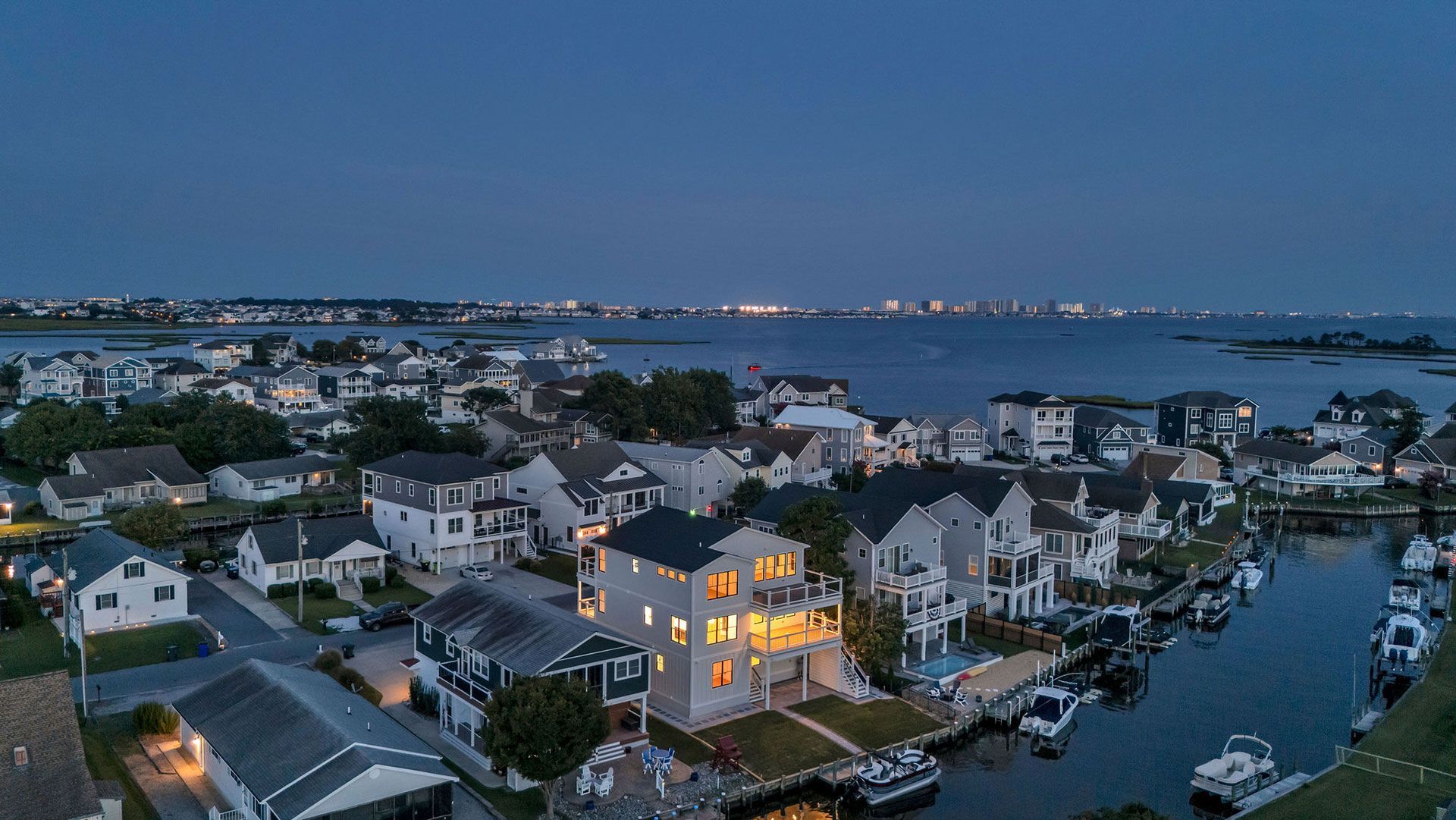 Aerial view of waterfront homes with lights on at dusk; boats docked on the water.