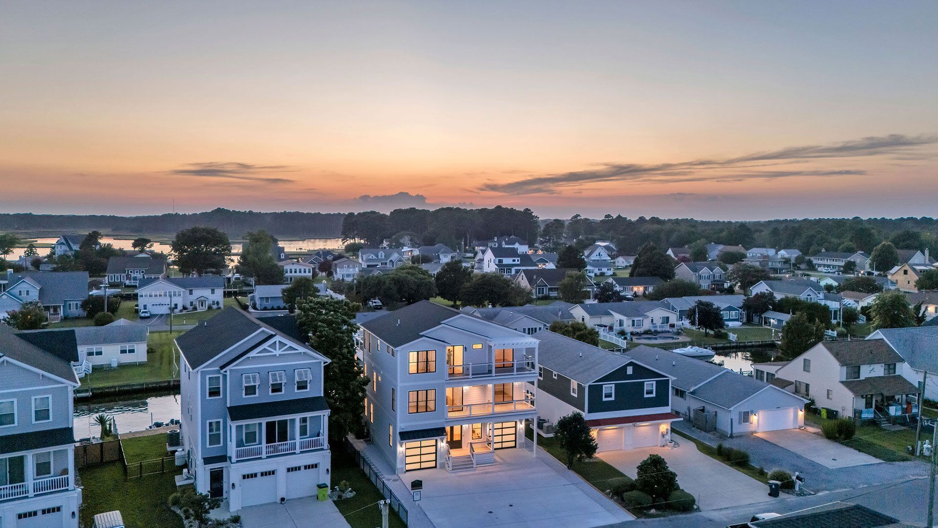 Aerial view of houses at dusk, waterfront location, orange sky, calm water, suburban area.