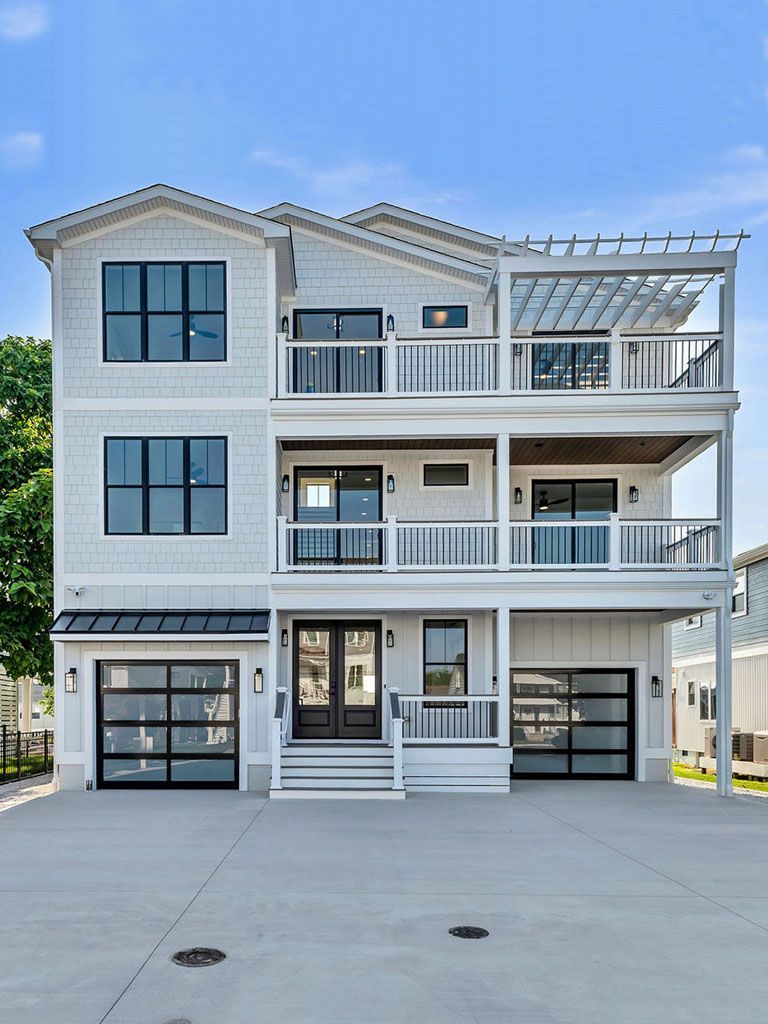Three-story light grey house with black window frames, balconies, and two glass garage doors; bright sky.