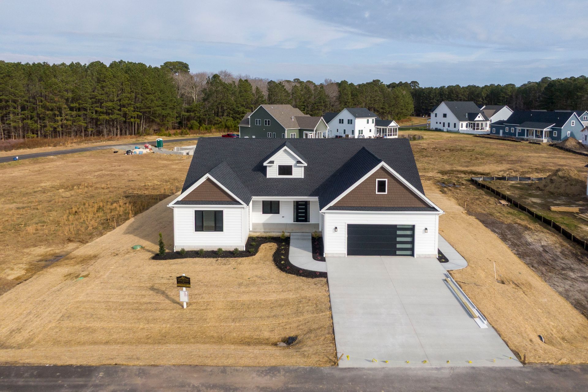 Aerial view of a white suburban house with a black roof and driveway, surrounded by bare land and nearby homes.