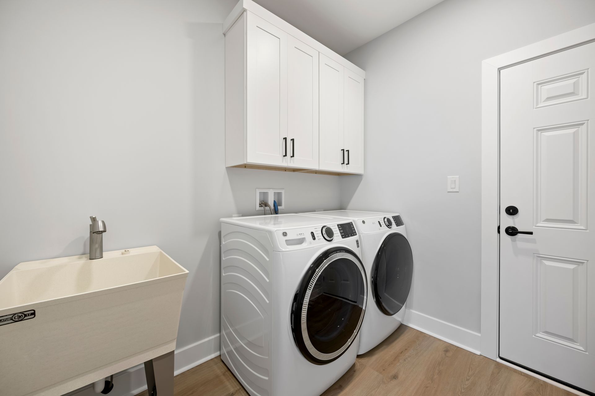Laundry room with white washer and dryer, utility sink, cabinets, and a closed door on wood flooring