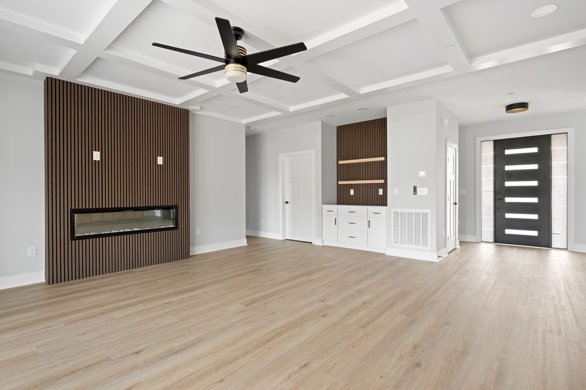 Modern empty living room with light wood floors, white coffered ceiling, ceiling fan, and dark wood accent wall.