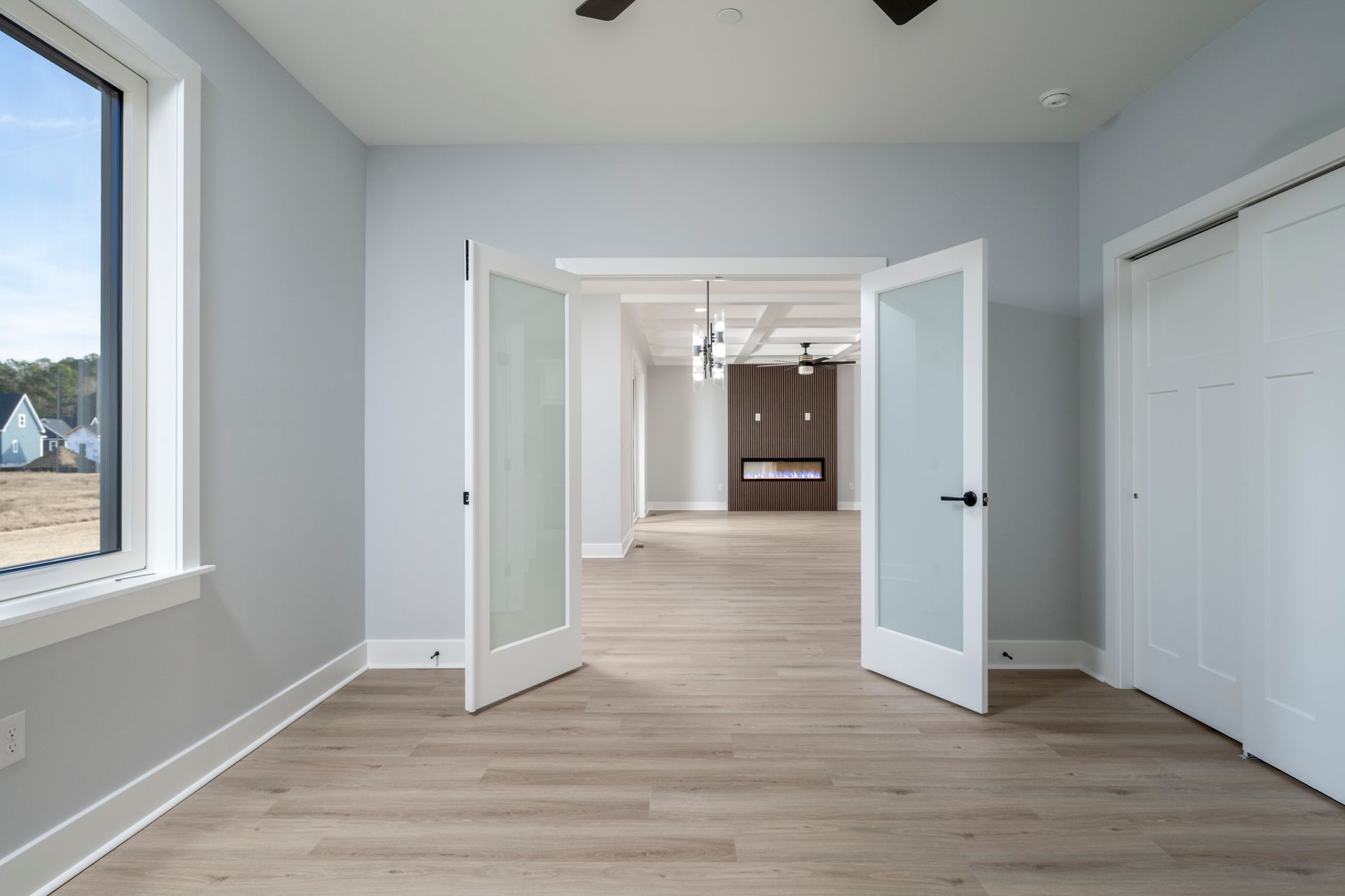 Bright empty room with light gray walls, white French doors, wood floors, and a large window.