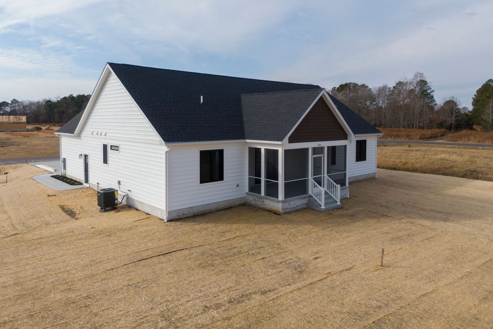 White house with black roof and small front porch on a bare lot under a cloudy sky
