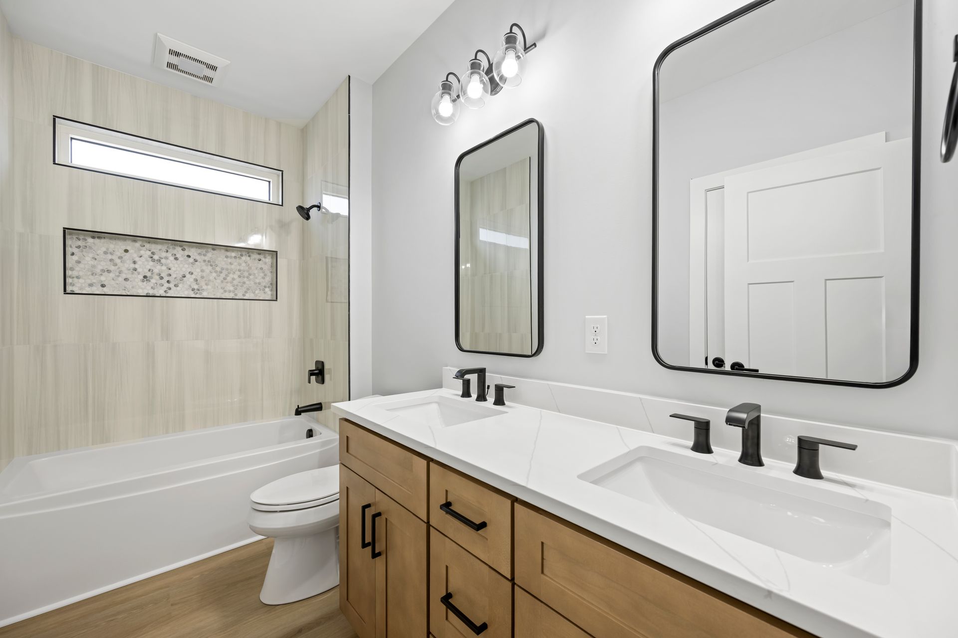 Bright modern bathroom with double vanity, large mirror, and white bathtub with tiled shower wall.