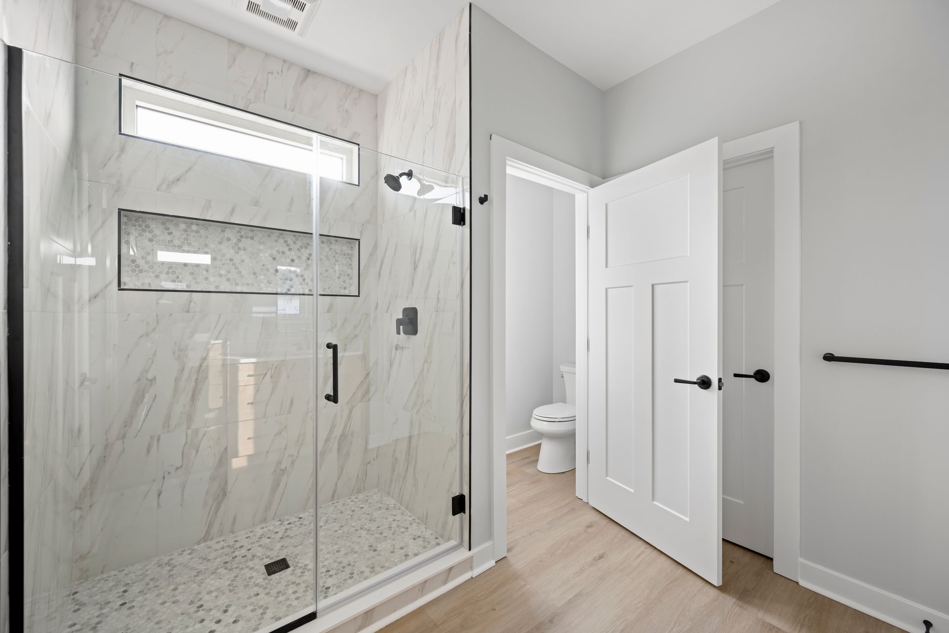 Bright modern bathroom with marble shower, glass door, and open white door to a toilet room