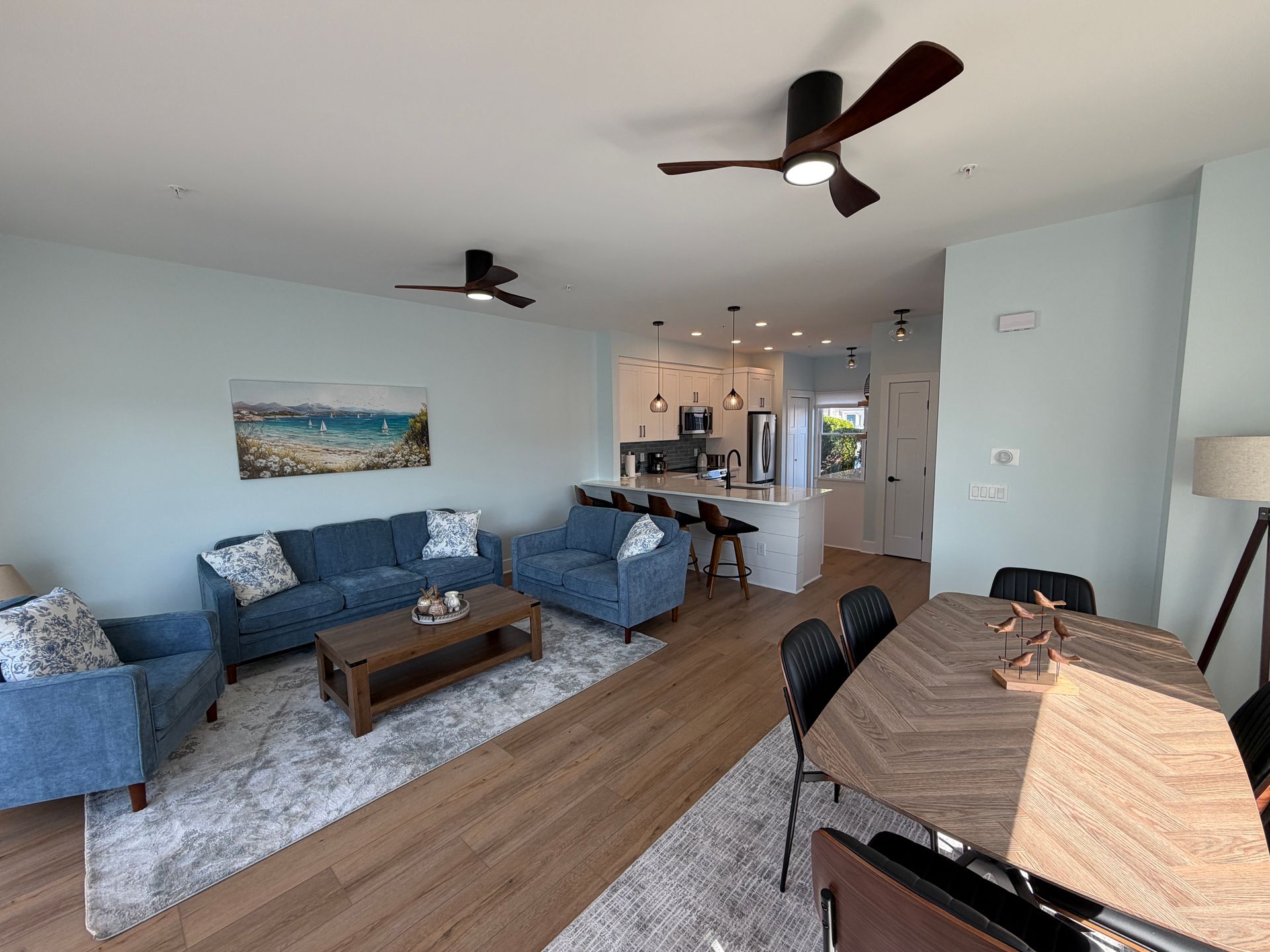 Empty modern living room with light wood floors, white walls, and ceiling fans, open to a kitchen.