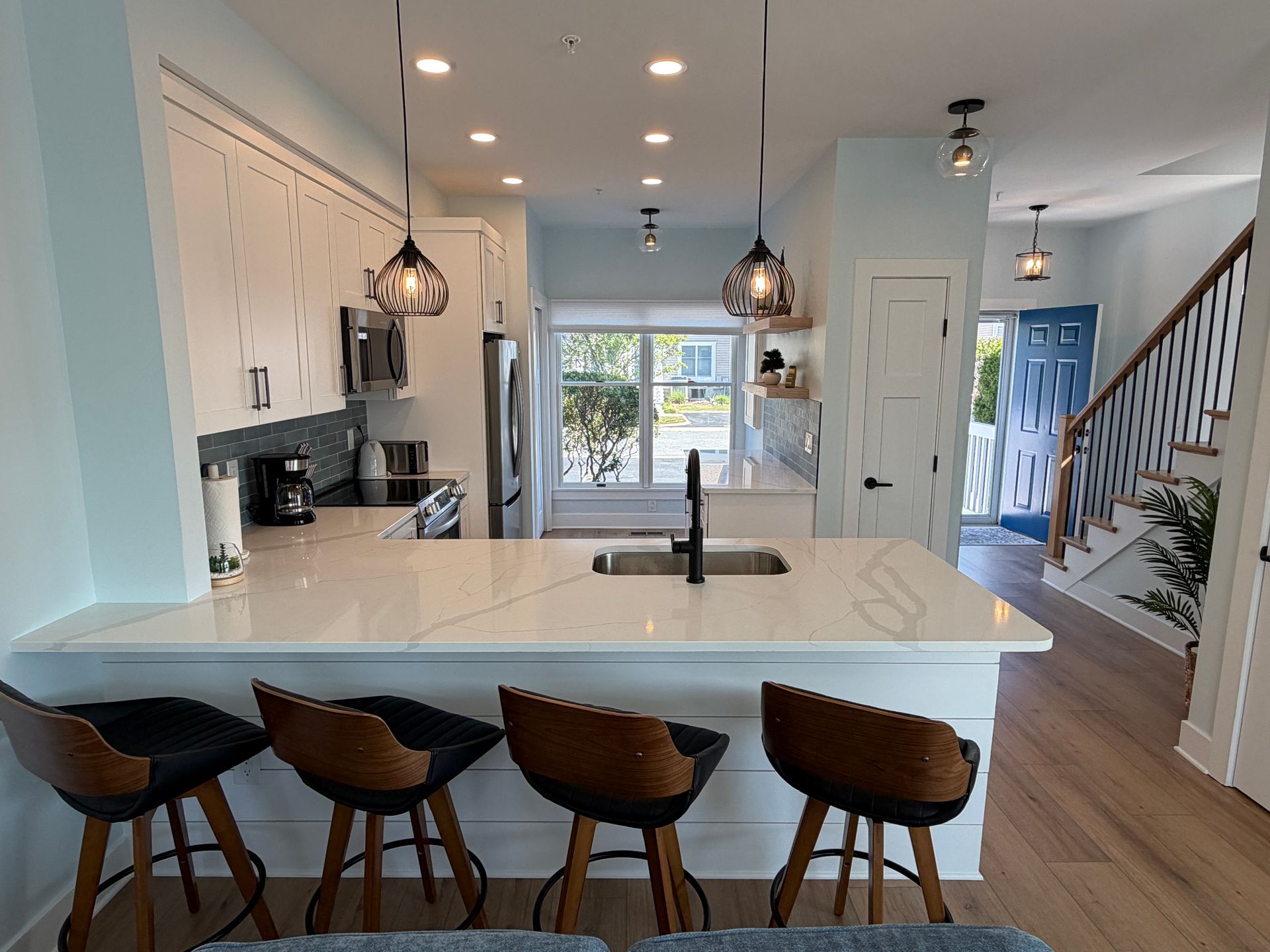 Bright white kitchen with island, pendant lights, and hardwood floor leading to stairs and a blue hallway.