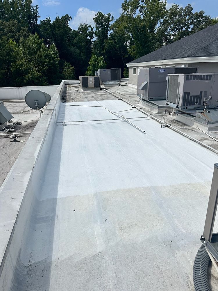 White-coated flat roof with HVAC units, antennas, and surrounding trees under a blue sky.