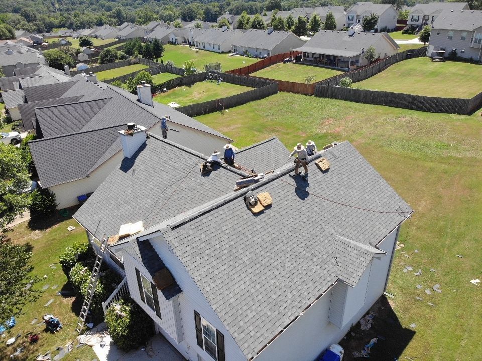 Workers on a roof installing shingles. Gray roof, green lawn, suburban setting.