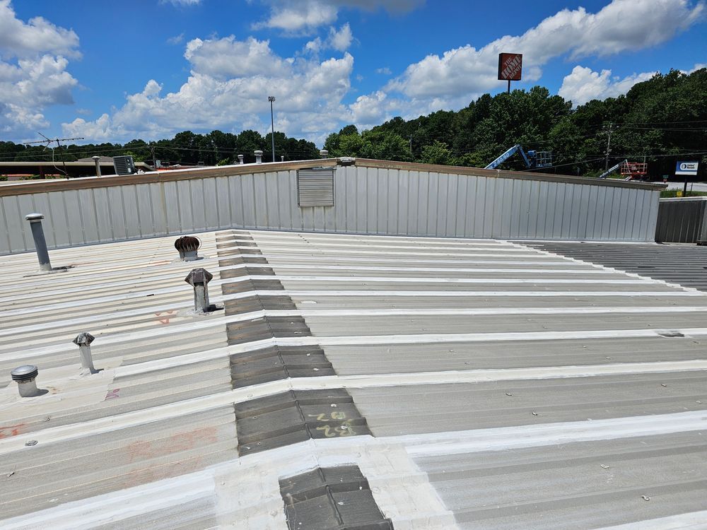 View of a gray commercial roof with vents, a low wall, and trees in the background under a blue sky.
