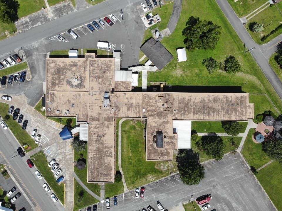 Overhead view of a school with a flat roof, surrounded by parking lots, grass, and trees.