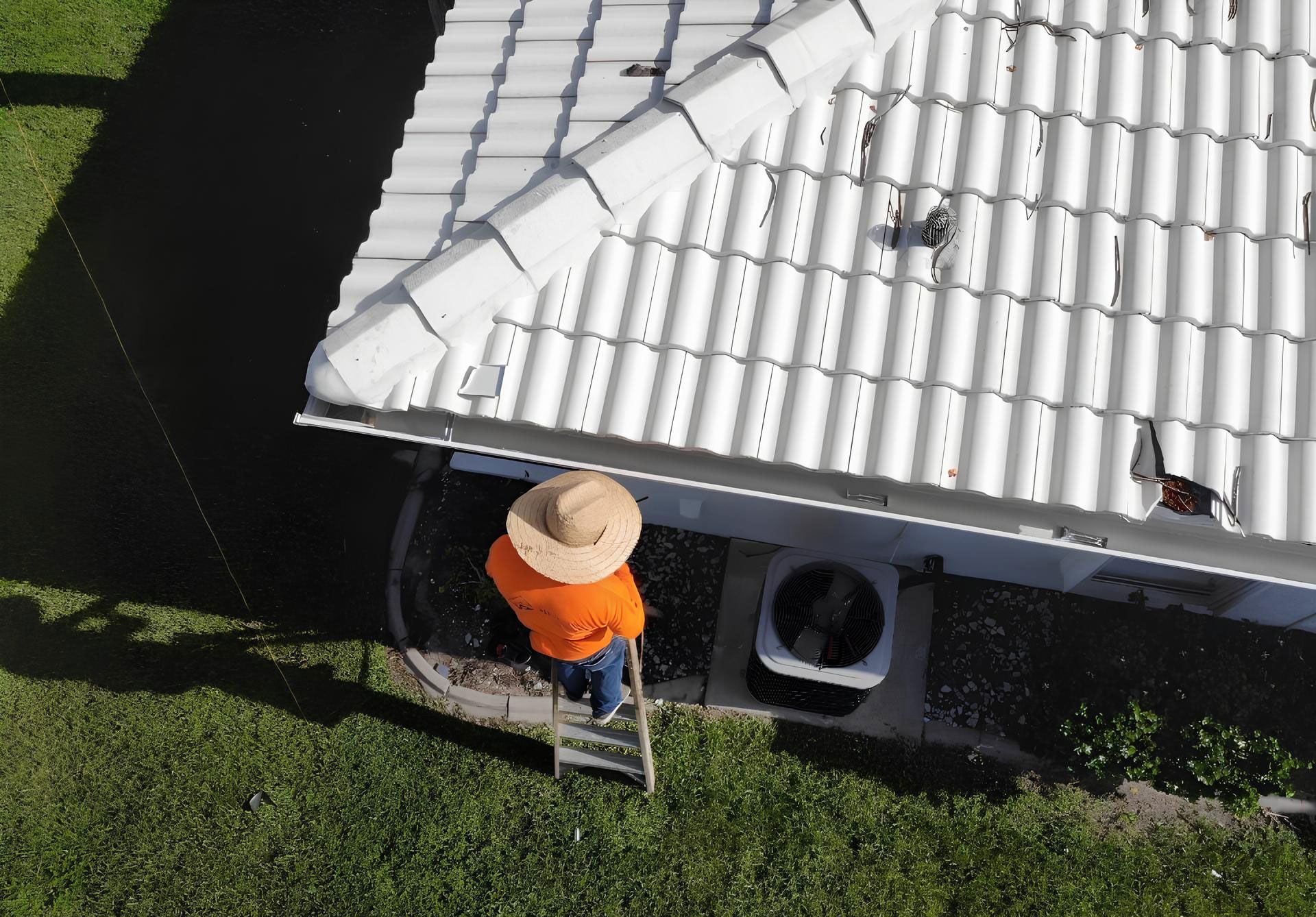 Person in an orange shirt and straw hat on a ladder, working on a white tiled roof.