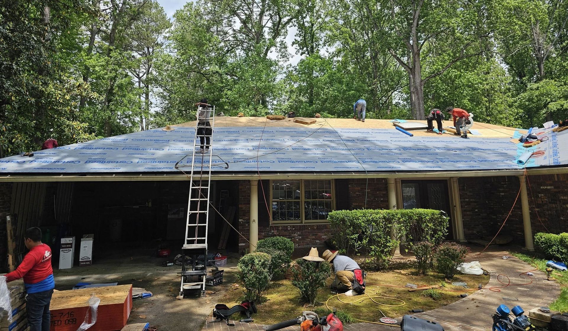 Roofers working on a residential roof, with materials, ladders, and tools visible; surrounded by trees.