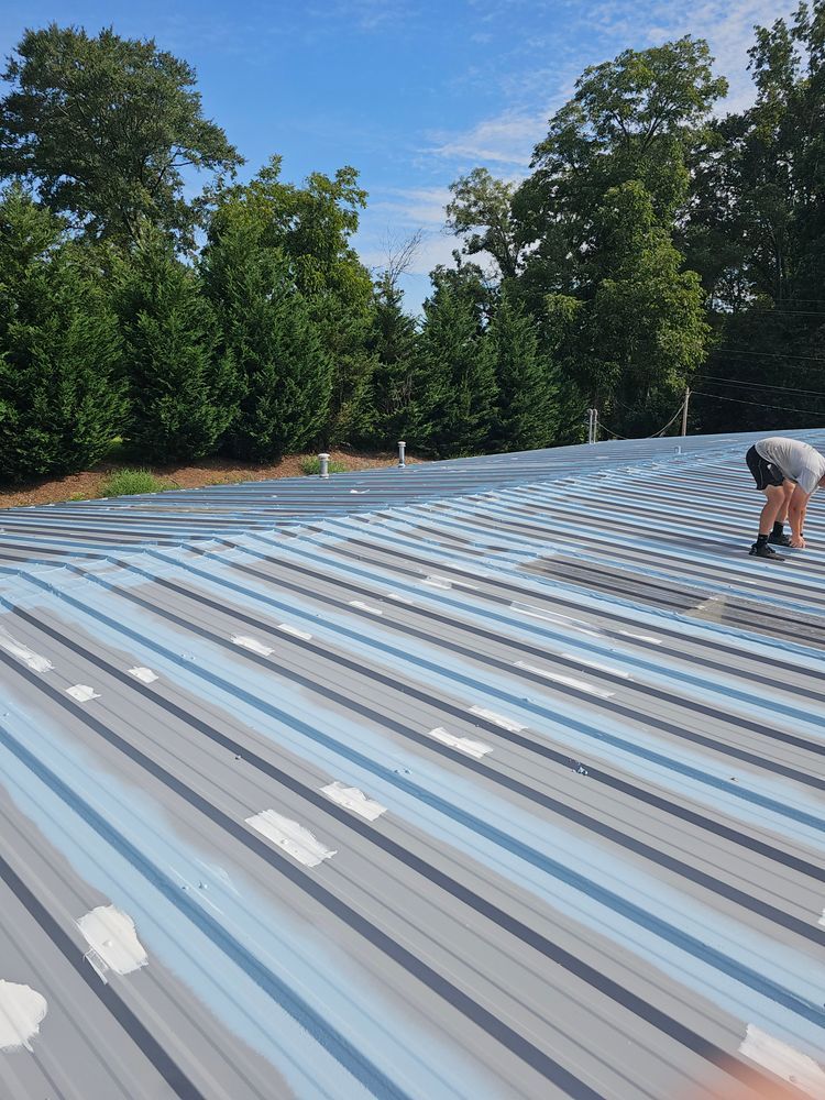 Person on a blue and gray metal roof, repairing paint. Trees and blue sky in the background.