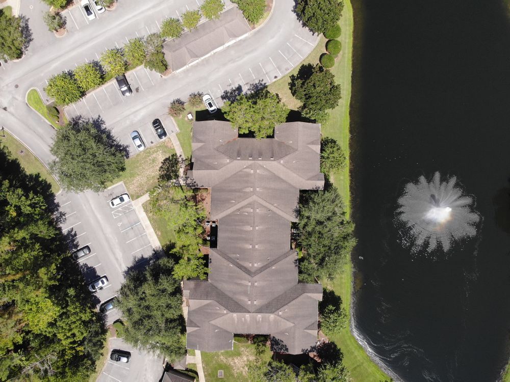 Aerial view of a building with a dark roof next to a pond with a fountain and parking lot.