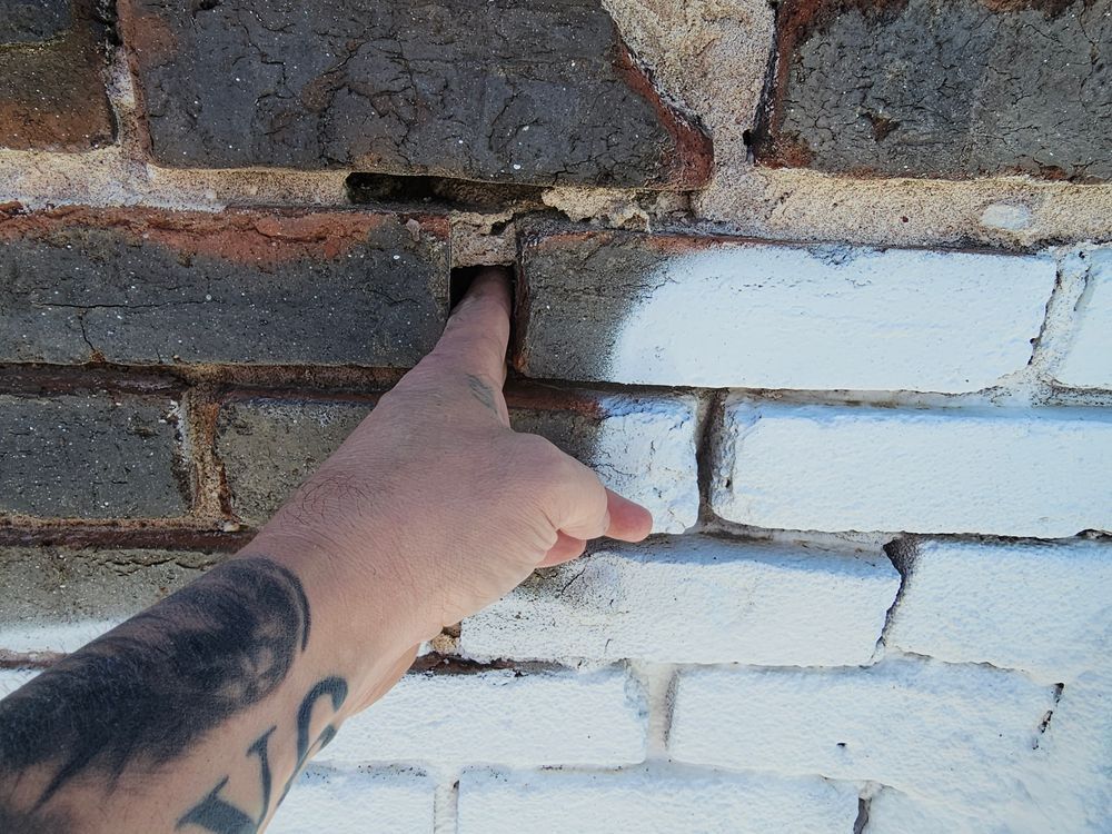 Hand points to a hole in brick wall with gray and white painted bricks.
