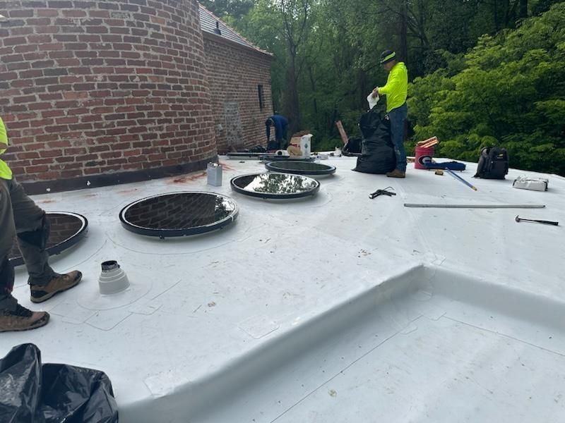 Workers on a white flat roof install or repair; a brick building and trees are in the background.