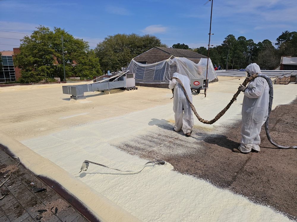 Two workers in protective suits spraying foam insulation on a roof. White foam covers the roof in sections.