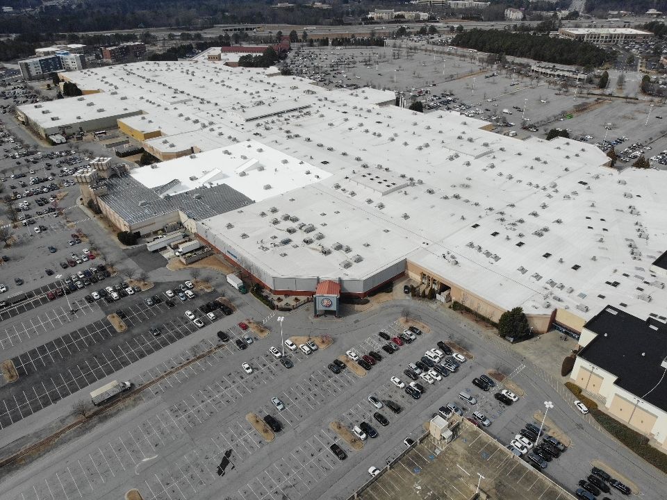 Aerial view of a large shopping mall with expansive parking lots, cars, and multiple roof sections.
