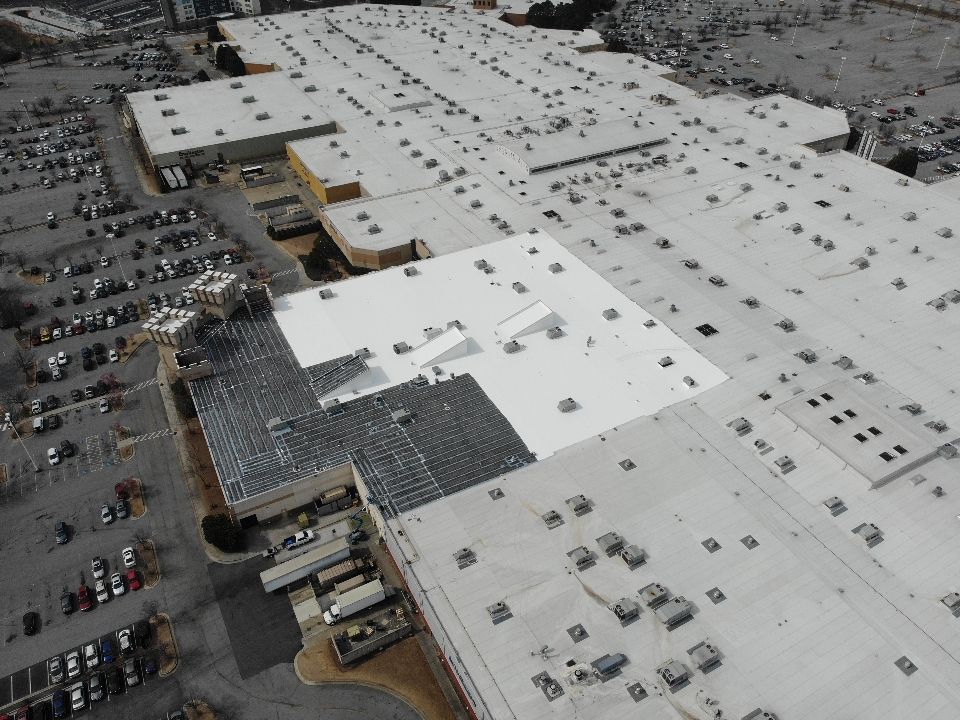 Aerial view of a large commercial building with a white roof and parking lot filled with cars.