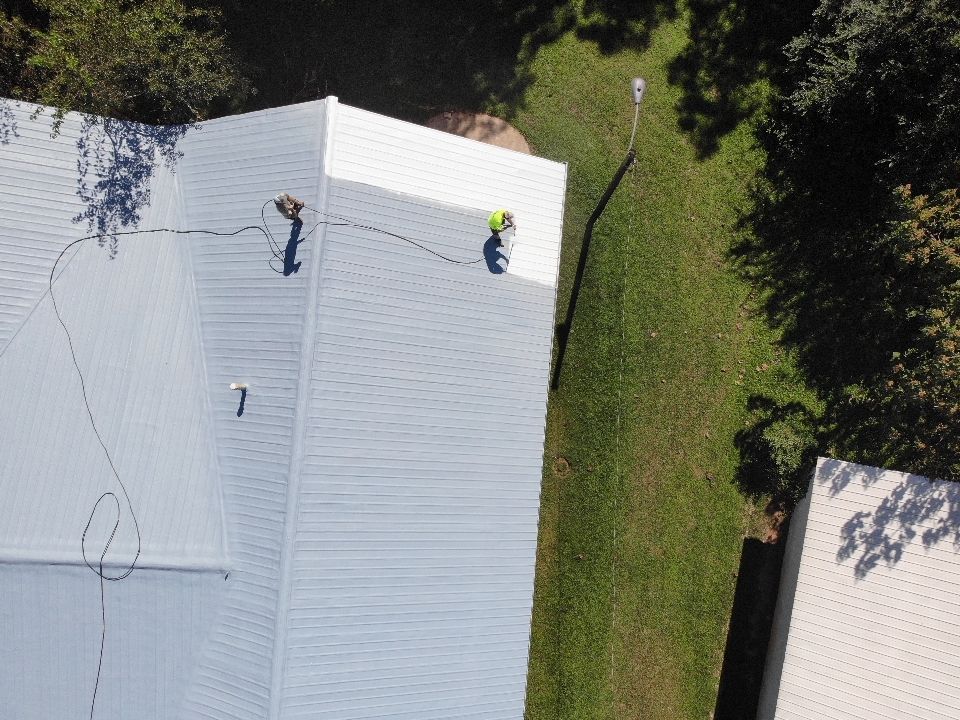 Workers on a metal roof, one in a yellow vest, near a grassy area.