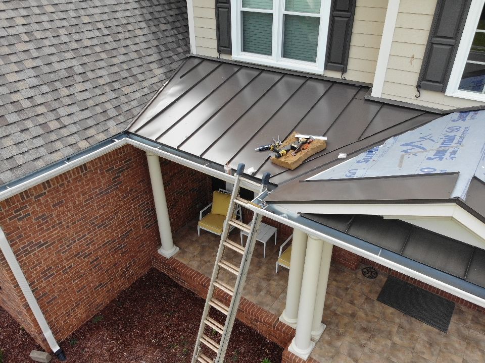 A brown metal roof being installed on a house with a brick exterior. A ladder leans against the porch.