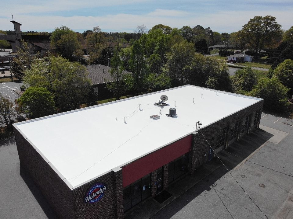 Aerial view of a commercial building with a white roof, surrounded by trees and residential homes.
