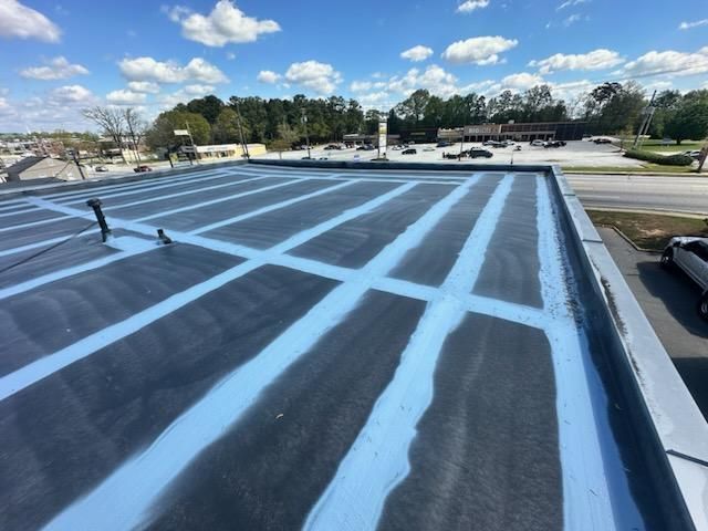 Flat commercial roof with dark surface and blue grid lines, trees and buildings in the background.