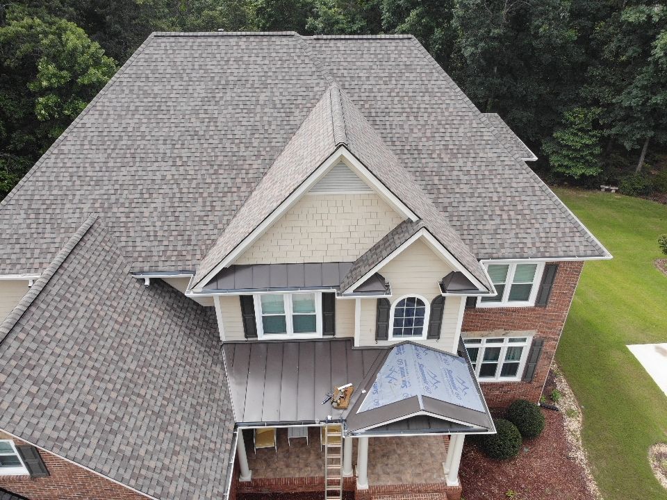 View of a house with a gray shingle roof and a metal roofed porch. The house is brick with several windows.