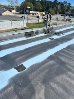A flat roof being repaired with light blue coating between dark gray sections. Buildings and cars are in the background.