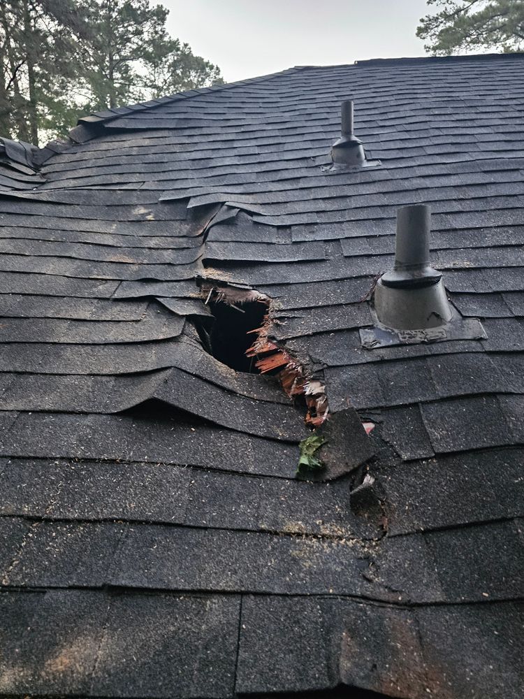 Damaged black shingle roof with a large hole revealing wood. Two vents are visible.