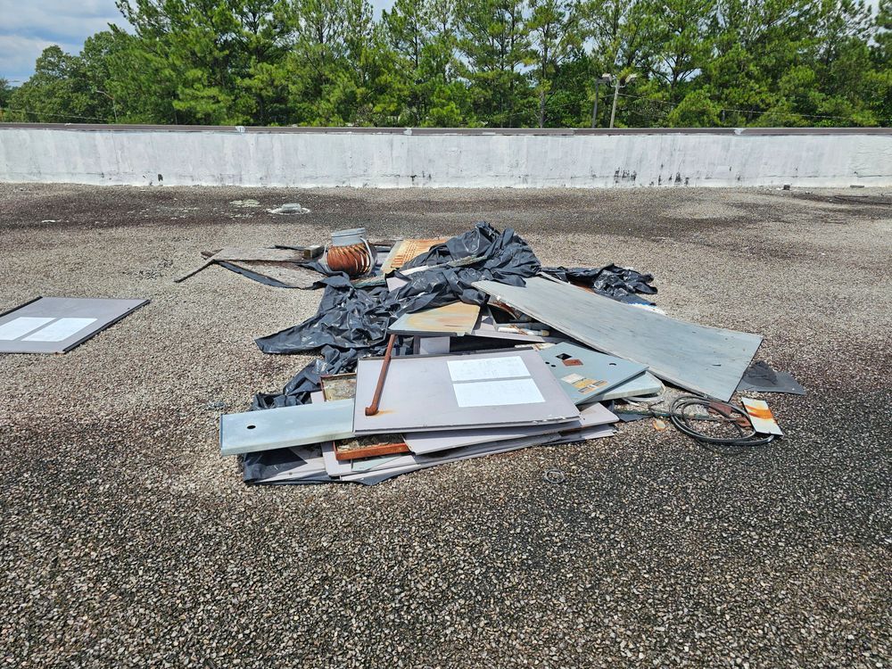 Pile of debris on a flat roof; includes metal, wood, and black plastic. Green trees and white wall in the background.