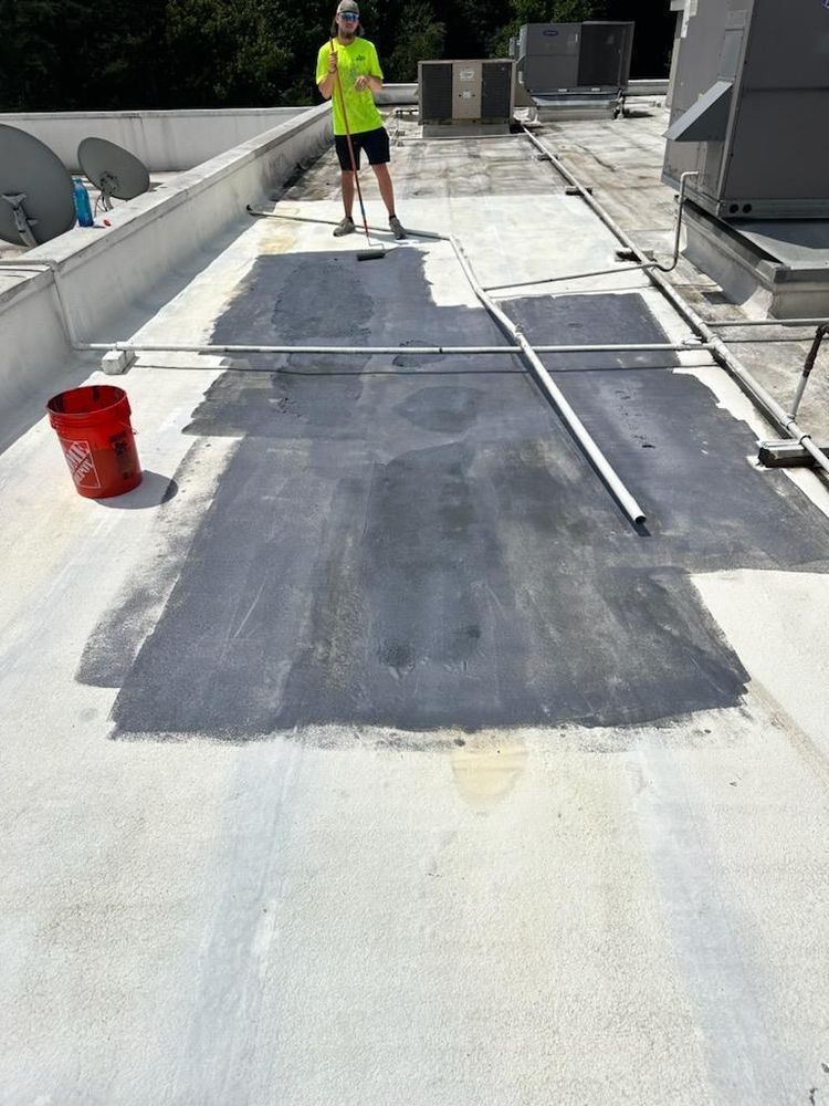 Person on a rooftop applies dark sealant over a large section of light-colored roofing material. Red bucket nearby.