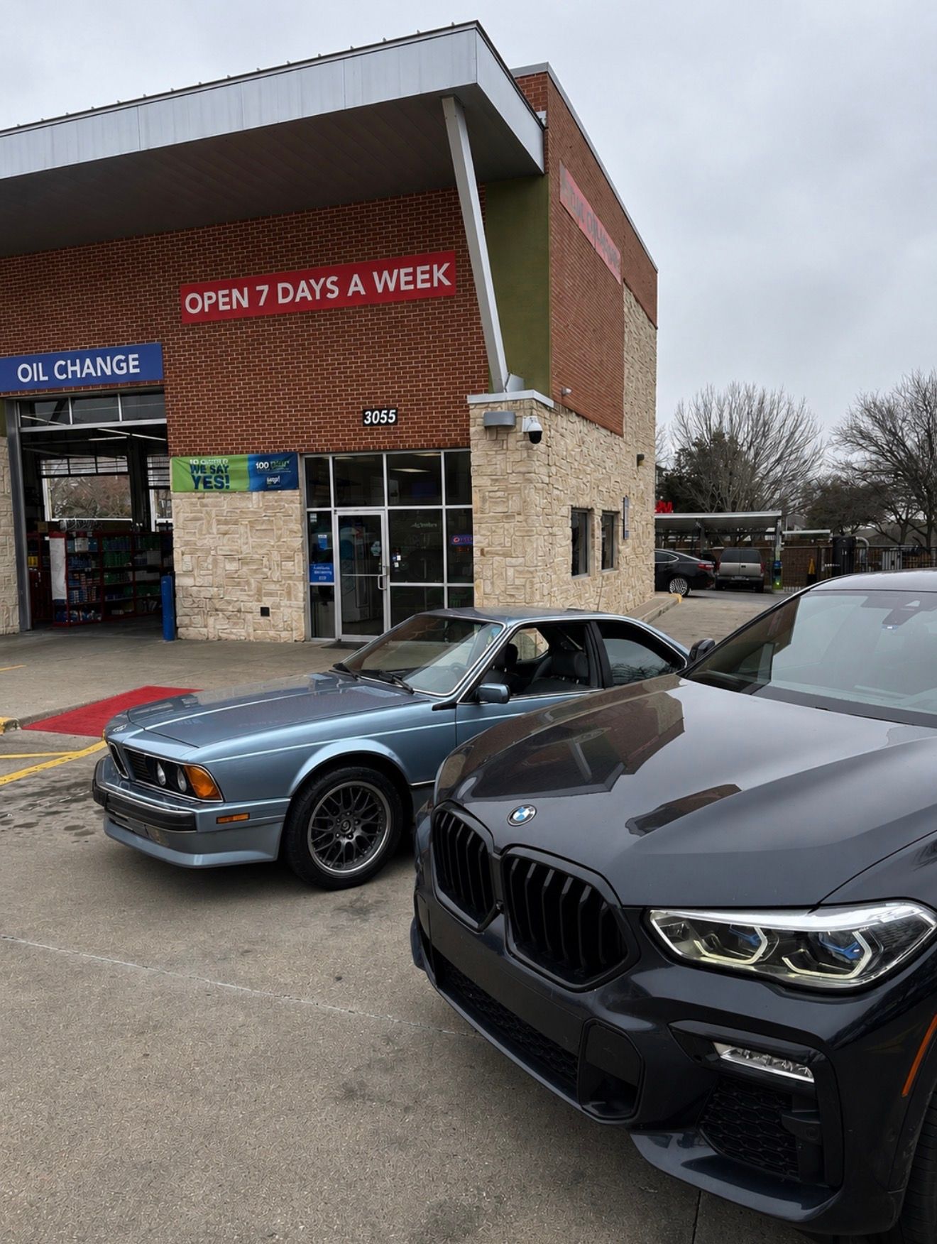 Two parked BMWs outside a brick storefront with a “OPEN 7 DAYS A WEEK” sign.