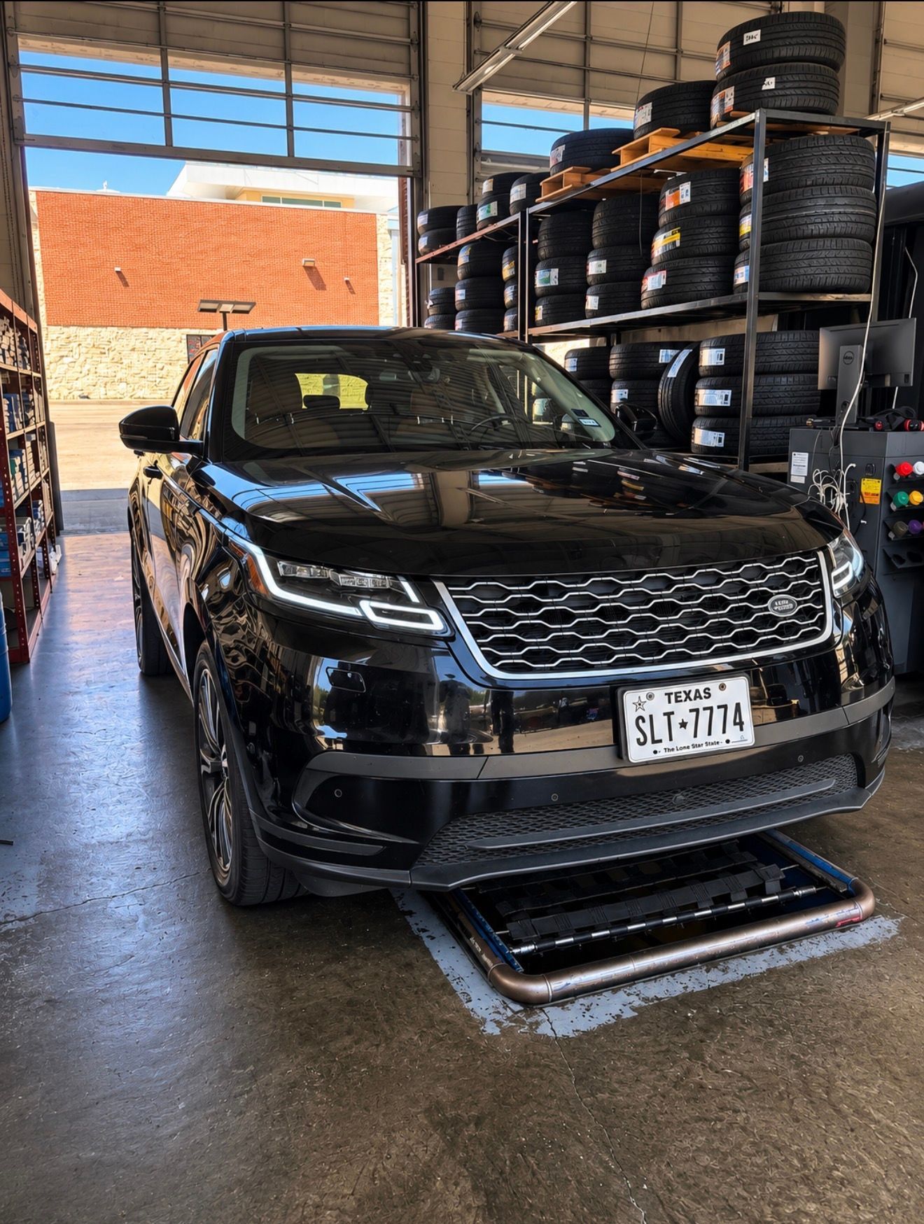 Black SUV in a tire shop bay, parked on a lift with stacked tires in the background.