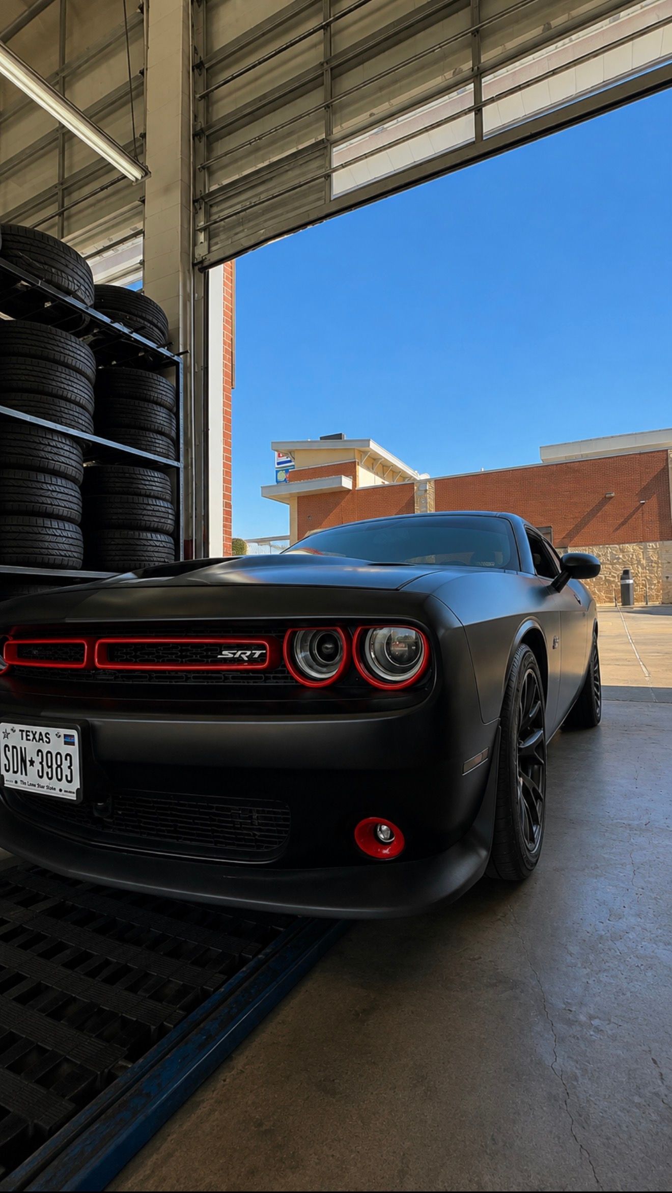 Black Dodge Challenger parked in a garage, with blue sky and buildings visible outside.