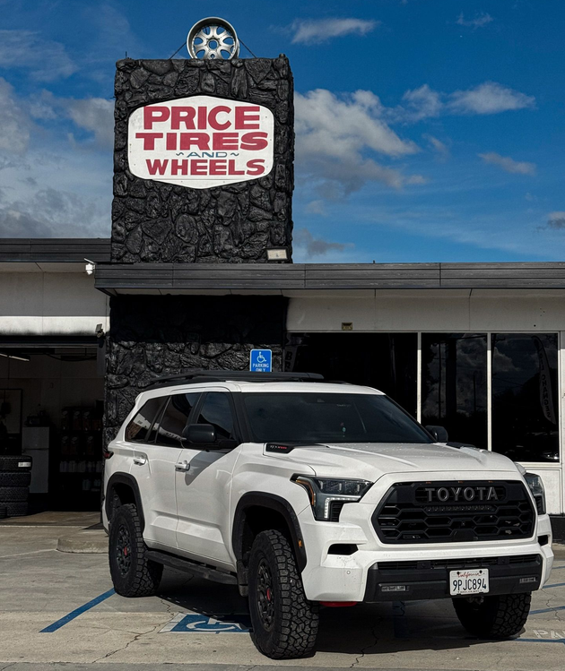 A white Toyota Sequoia parked in front of a Price Tires and Wheels shop with a dark stone facade.