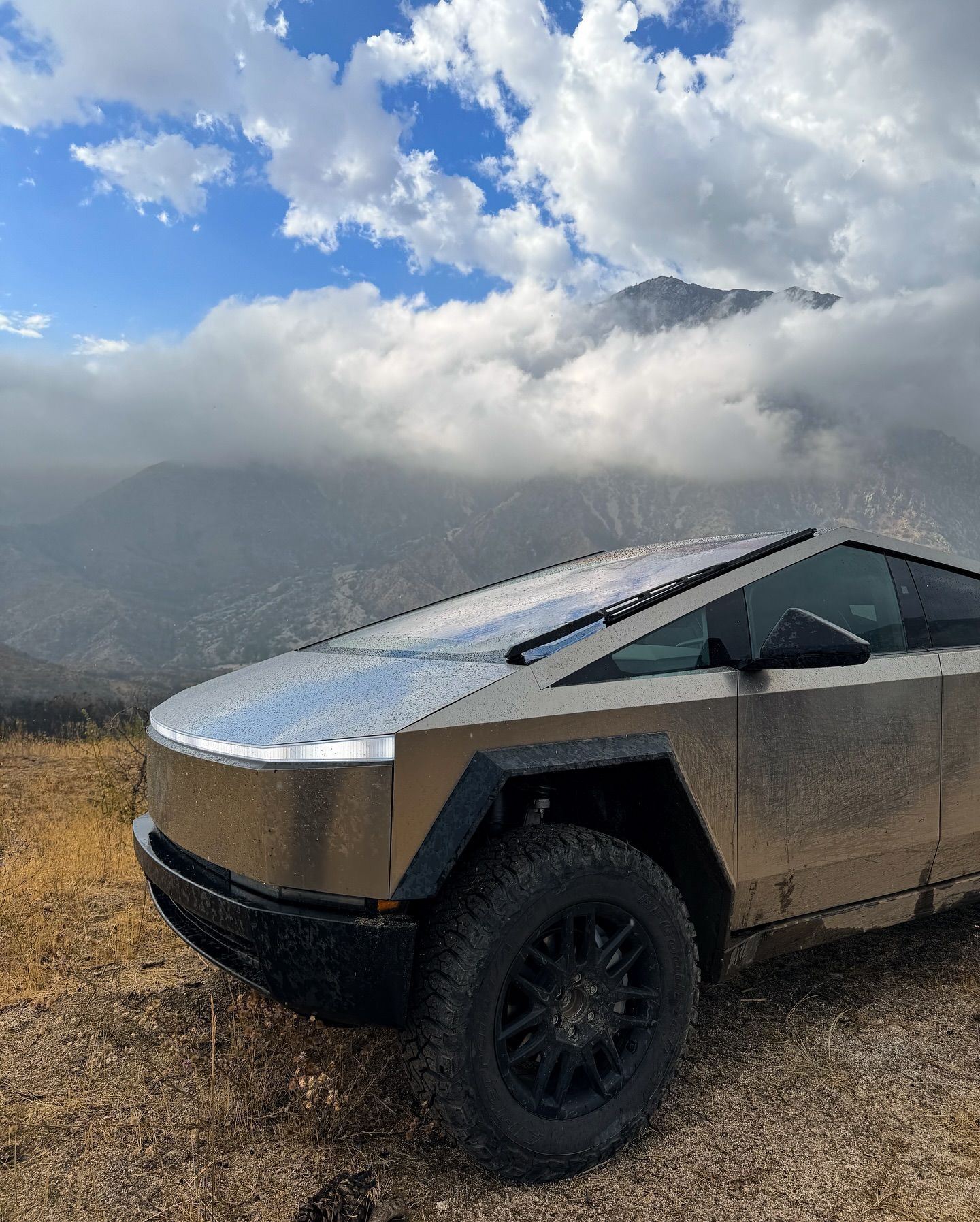 A Tesla Cybertruck parked on a dirt path with rugged mountains and a cloudy sky in the background.