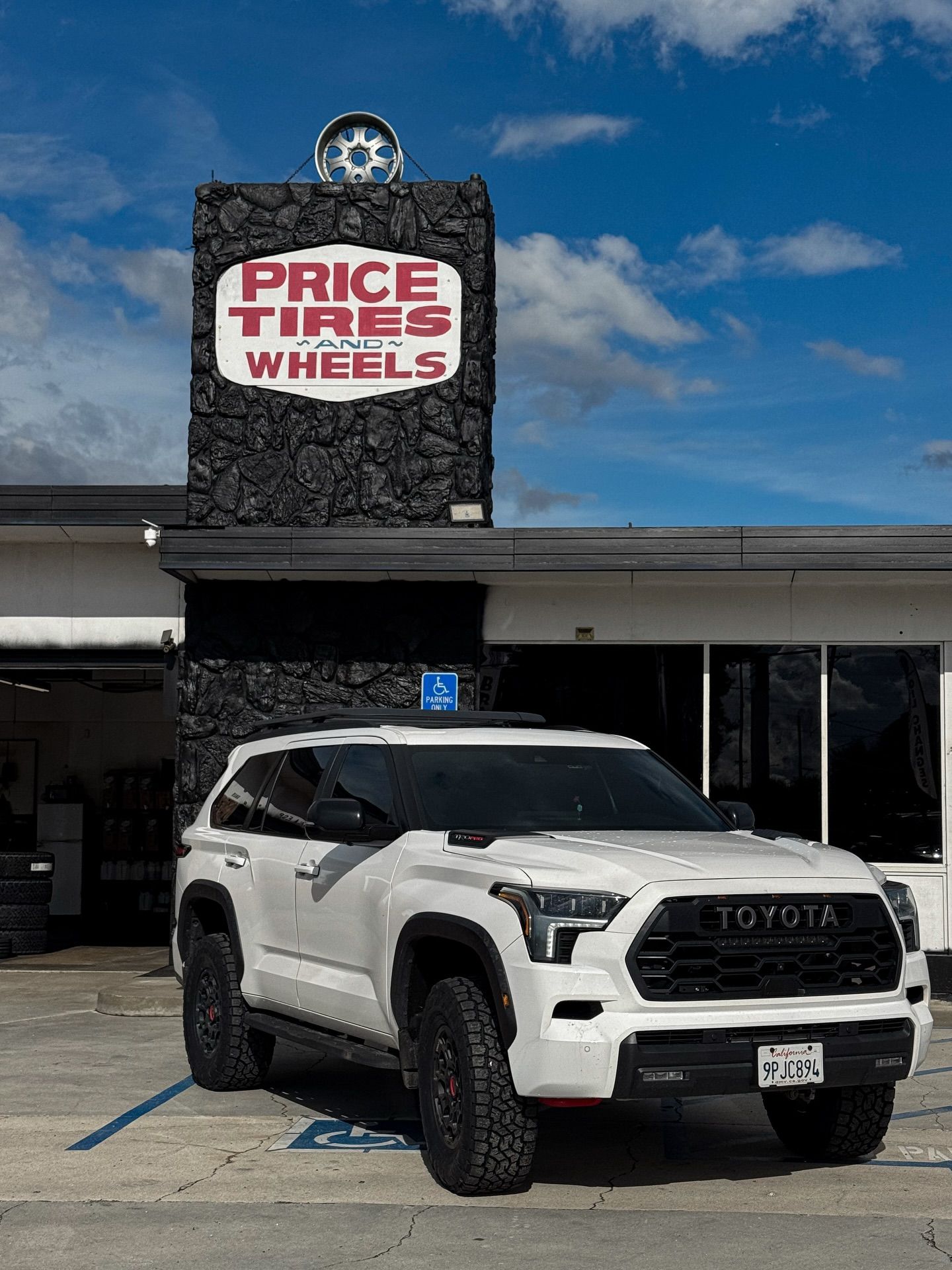 A white Toyota SUV parked in front of a Price Tires and Wheels shop with a stone facade and a wheel mounted on top.