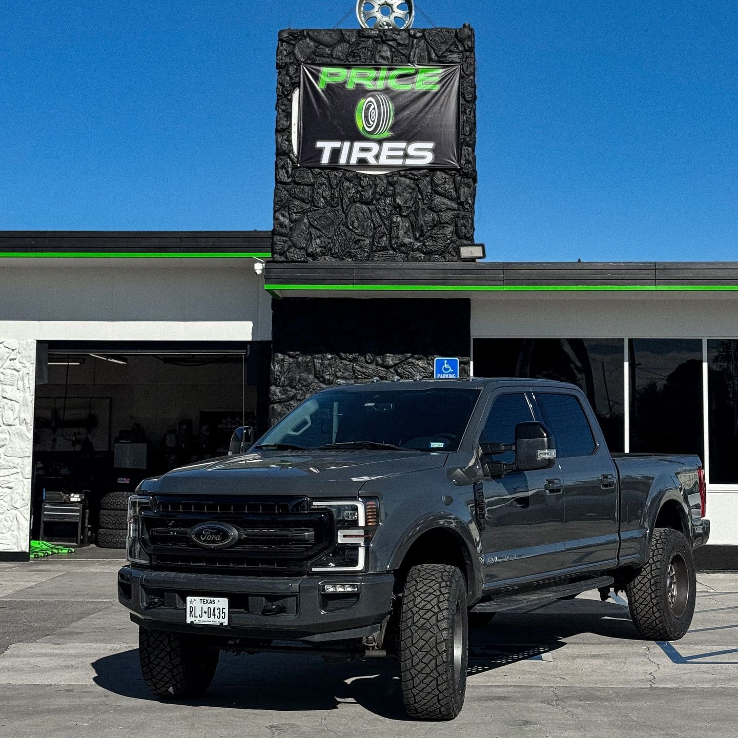 A gray Ford pickup truck parked in front of a Price Tires building with a black stone facade and a green and white sign.