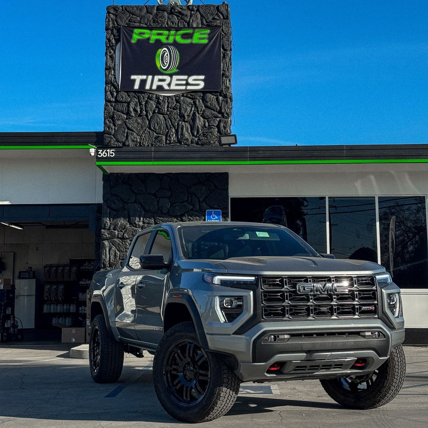 A gray GMC Canyon pickup truck parked in front of a Price Tires building with a black stone facade.