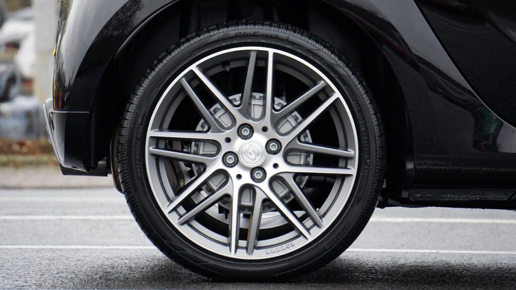 A close-up shot of a modern, multi-spoke silver alloy car wheel with a black tire on an asphalt surface.