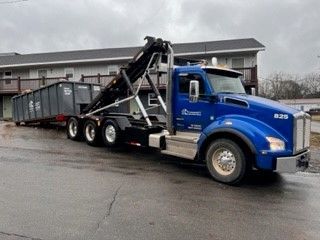 A blue dump truck is carrying a dumpster on a trailer.