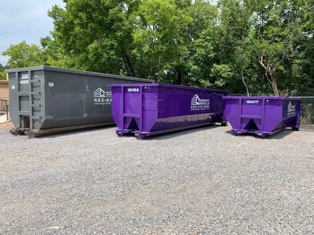 Three purple dumpsters are parked next to each other in a gravel lot.