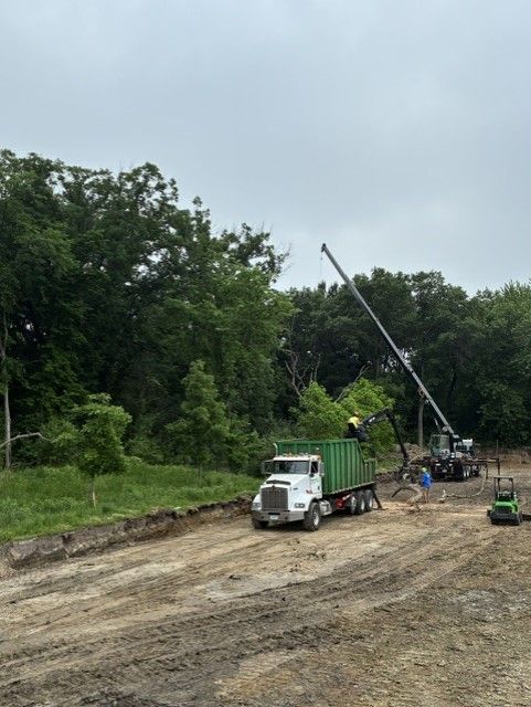 Construction site with a white truck, green crane, and workers on a dirt road near trees
