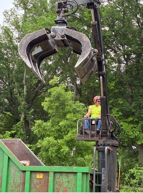 Large claw attachment lifts debris beside a worker in a neon safety vest at a green industrial site.